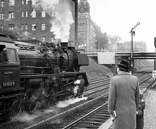 Ein Mann und ein Kind stehen auf einem Bahnsteig und schauen der dampfenden Reko-Lokomotive nach, die an ihnen vorbei rauscht.
