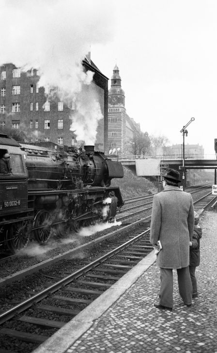 Ein Mann und ein Kind stehen auf einem Bahnsteig und schauen der dampfenden Reko-Lokomotive nach, die an ihnen vorbei rauscht.