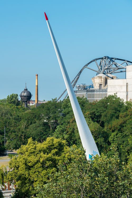 A large, white rotor blade rises vertically out of green treetops. In the background, the curved supporting girders of a roof structure, a water tower, and a chimney can be seen.