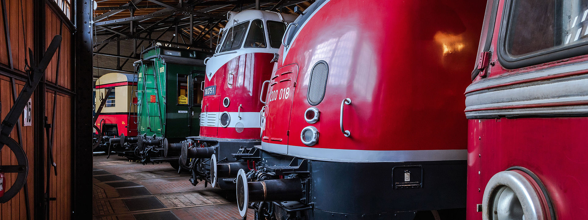 Blick in die Schienenverkehrs-Ausstellung im historischen Lokschuppen: Drei rote Dieselloks und zwei Abteilwagen stehen auf Schienen nebeneinander aufgereiht.