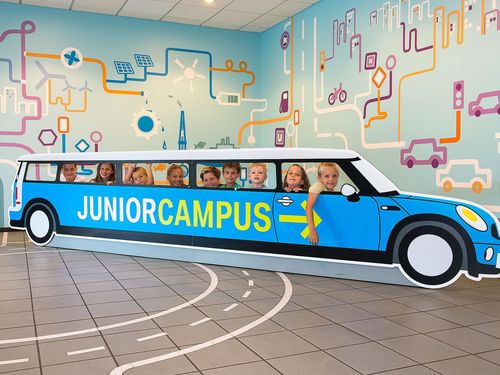 Neun Kinder schauen durch die Seitenfenster eines etwa sechs Meter langen Aufstellers aus Pappe in Form eines Autos. Auf der Seite ist der Schriftzug „Junior Campus“ in grün auf blauem Grund angebracht. Auf dem Boden sind Fahrbahnmarkierungen angedeutet.