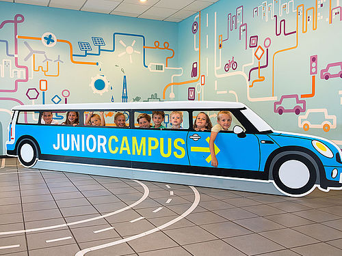 Neun Kinder schauen durch die Seitenfenster eines etwa sechs Meter langen Aufstellers aus Pappe in Form eines Autos. Auf der Seite ist der Schriftzug „Junior Campus“ in grün auf blauem Grund angebracht. Auf dem Boden sind Fahrbahnmarkierungen angedeutet.