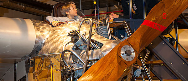 A young boy sits in the cockpit of a historical airplane. A man kneels on a ladder, at eye level with the boy.