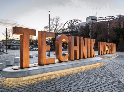 Auf einem gepflasterten Platz sieht man eine aus Metallbuchstaben erstellte Wortskulptur, die sich „Technik Museum“ liest. Im Hintergrund die Silhouette des Technikmuseums. 