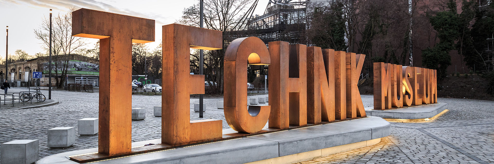 Auf einem gepflasterten Platz sieht man eine aus Metallbuchstaben erstellte Wortskulptur, die sich „Technik Museum“ liest. Im Hintergrund die Silhouette des Technikmuseums. 