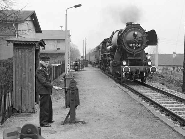  A black and white photograph: An elderly gatekeeper in uniform is standing on a train platform set in a rural area. A steam locomotive is approaching the scene from the background.