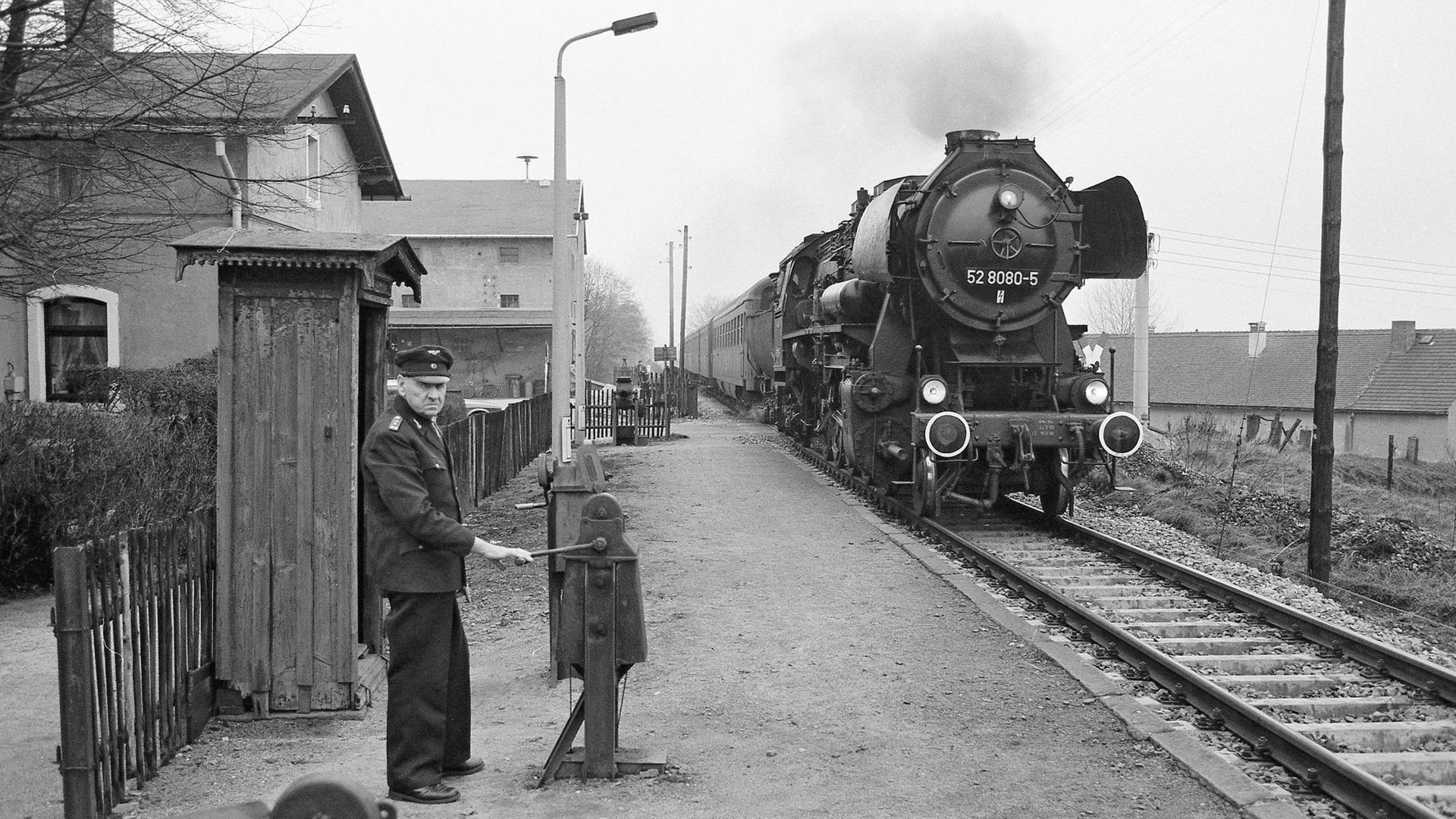 Eine Schwarzweiß-Fotografie: Auf einem Bahnsteig in ländlicher Umgebung steht ein älterer Schrankenkwärter in Uniform. Von hinten nähert sich eine Dampflokomotive.