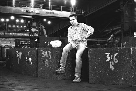Der Fotograf Günter Krawutschke sitzt mit kariertem Hemd, Jeans und Turnschuhen auf einem Betonblock in einer Fabrikhalle. Neben ihm liegt ein Bauhelm.