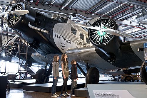 Drei Kinder stehen vor einem sehr großen Flugzeug in der Ausstellung und betrachten das Triebwerk.