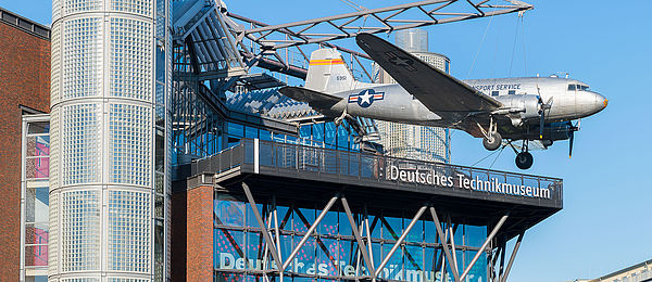 A modern building with a glass front. An airplane is suspended from a steel contruction on the roof.