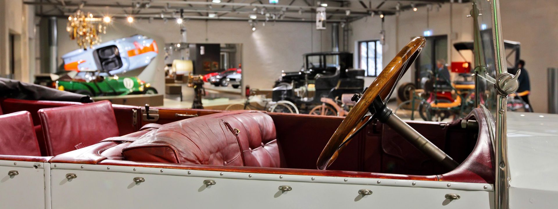 Blick über das offene Verdeck eines historischen weißen Cabriolets in die Straßenverkehr-Ausstellung. Sitze und Verkleidung sind mit dunkelrotem Leder bezogen, das Lenkrad ist aus dunklem Holz. 