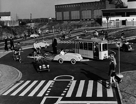 Schwarz-Weiß-Aufnahme, Kinder auf einem Verkehrsparcours in unterschiedlichen Fahrzeugen, im Vordergrund überqueren zwei Kinder einen Zebrastreifen.
