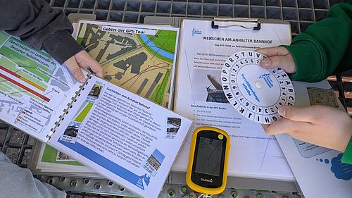 Zusehen sind die Informationsmaterialien für die GPS-Tour, eine Entschlüsselungsscheibe und ein GPS-Gerät.