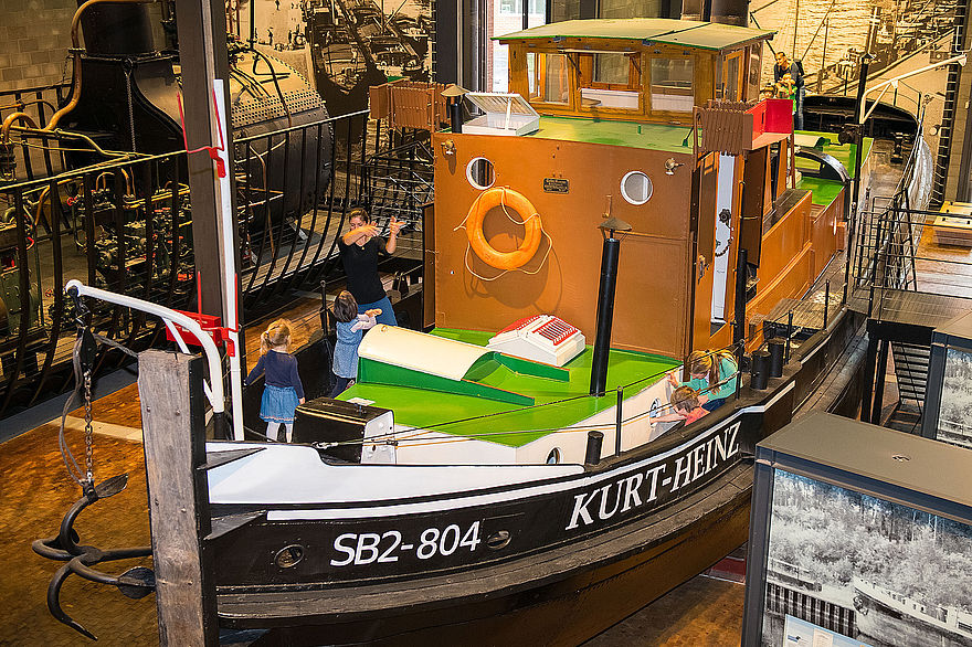 Ein kleines, historisches Dampfschiff steht in der Schifffahrt-Ausstellung. Mehrere Personen stehen auf dem Vorderdeck. Seitlich ist der Schriftzug „SB2 -804“ und „Kurt-Heinz“ zu lesen. 