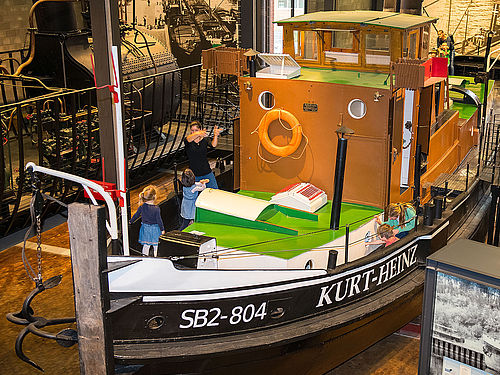 Ein kleines, historisches Dampfschiff steht in der Schifffahrt-Ausstellung. Mehrere Personen stehen auf dem Vorderdeck. Seitlich ist der Schriftzug „SB2 -804“ und „Kurt-Heinz“ zu lesen. 