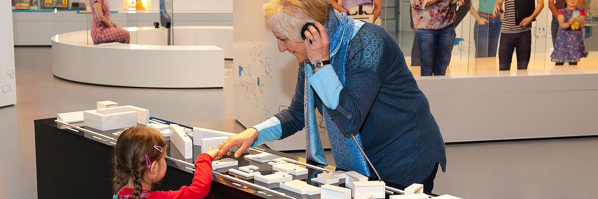 Eine ältere Frau und ein Kind stehen sich an einem Ausstellungstisch gegenüber. Auf dem Tisch befindet sich ein Übersichtsplan der Ausstellung zum Tasten. Die Frau hält sich den Hörer einer Hörstation ans Ohr. Gemeinsam tasten die beiden auf dem Plan.