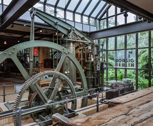 View of the flywheel of a green steam engine. It is partially sunk in the floor and is surrounded by an iron security barrier. Light enters the exhibition space through a glass roof and a wall of windows. There is an illuminated sign in front of the windows with the words “Borsig Berlin” on it.