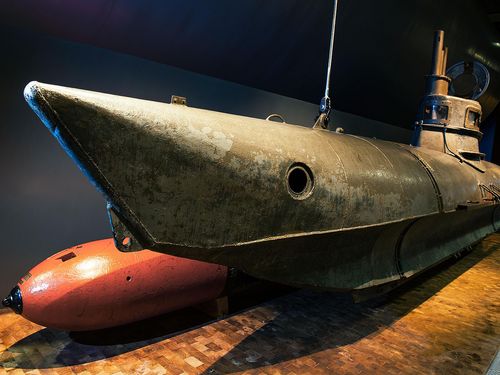 Schrägansicht des Einmann-U-Boots Bieber. Das U-Boot ist etwa neun Meter lang, verfügt über einen kleinen Turm mit Aussichtsfenstern und besteht aus grauem Metall. Die Nase des U-Boots ist nach oben gebogen. Dahinter liegt ein roter Torpedo.