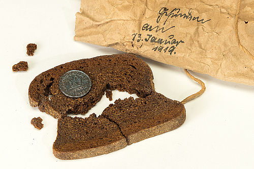 Das Bild zeigt eine hundert Jahre alte Scheibe Brot mit Münze. Das Geldstück wurde vermutlich für eine Hochzeit oder ähnliches eingebacken.
