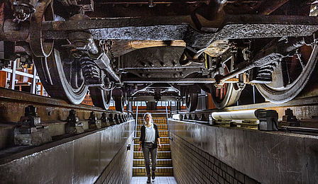 A young woman stands in a narrow passageway under a historical locomotive. The underside of the locomotive, with its wheels and chassis, can be seen above her.