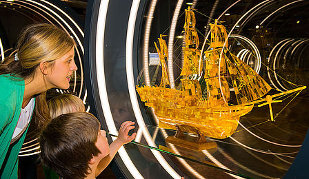 Eine Frau und zwei Jungen betrachten ein glänzendes Schiffsmodell aus Bernstein in einer runden, beleuchteten Vitrine. 
