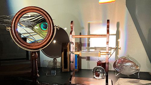 Museales Vergrößerungsglas mit einer Pfauenfeder in einem runden Holzrahmen, optischen Experimentiergeräten und Glasobjekten. Lichtbrechungen erzeugen farbenfrohe Regenbogenreflexe an der Wand.