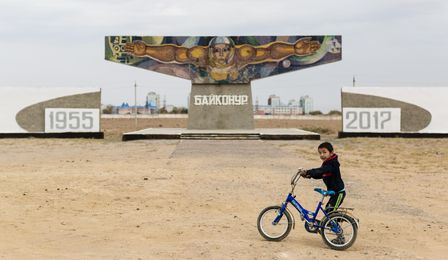 A small boy is pushing a bicycle across a sandy open space. In the background is a large monument showing the bust of a cosmonaut with his arms spread wide.