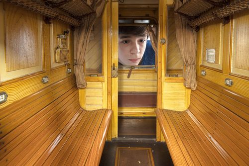 A boy looking into the window of a train model. In the foreground you can see the wooden benches of the 3rd class compartment.