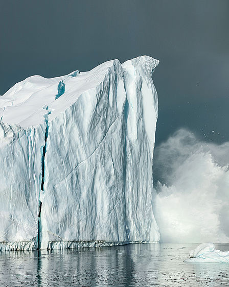 Von einem großen, schwimmenden Eisberg bricht ein Stück Eis ab, Wasser spritzt in einer großen Sprühwolke hoch. 