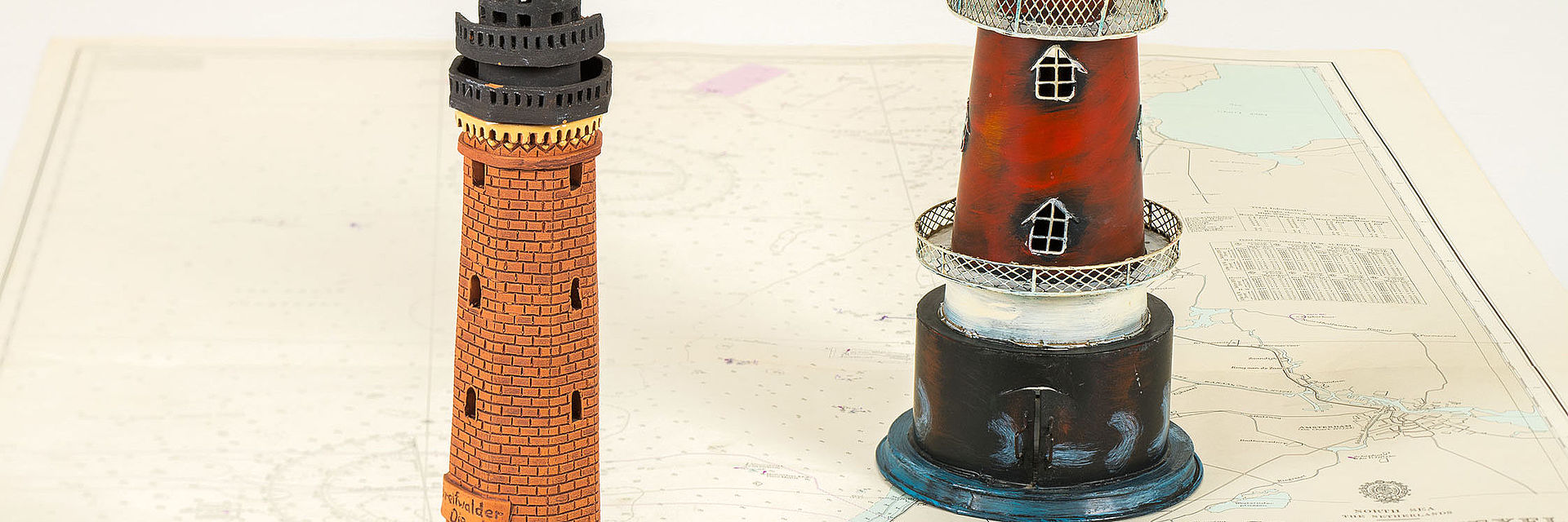 Auf einer Seekarte stehen das Modell eines Leuchtturms aus Backstein und das eines rot-weißen Fantasie-Leuchtturms.