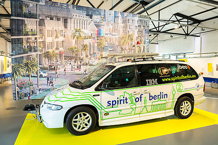 Weißes Automobil mit grünem Aufdruck von Berliner Wahrzeichen und einem blauen Schriftzug "spirit of berlin", dahinter hängt ein Banner mit einer Abbildung zur Mobilität der Zukunft.