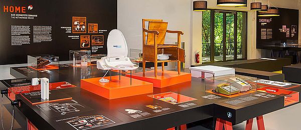 The HOME exhibit is a large table featuring objects in display cases, table-top media stations, texts, and graphics. A small, tangerine-colored model of a house is suspended above the table.