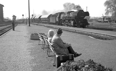 Ein Mann und eine Frau sitzen auf einer Bank auf einem menschenleeren Bahnsteig und schauen einem vorbeifahrenden Güterzug entgegen. Neben ihnen steht ein Koffer. 