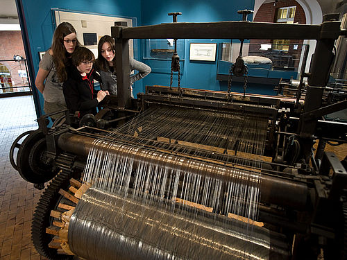 Three children look at a large, historical wire weaving loom in the exhibition.
