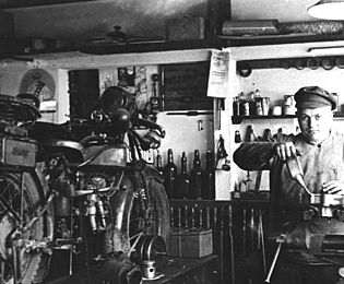Das Schwarz-Weiss-Foto ziegt links ein auf einem Tisch stehendes Motorrad. Rechts daneben schaut und Mann mit Mütze in die Kamera und ölt mithilfe einer Ölkanne ein Motorenteil. 