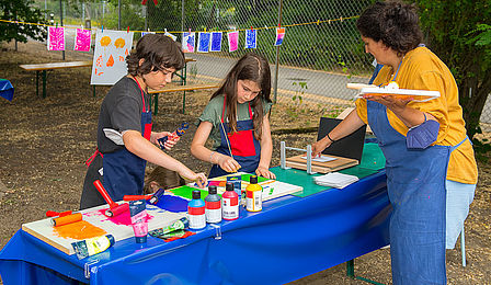 Zwei Kinder und eine Frau stehen an einem Tisch und drucken mit bunten Farben Pflanzenmotive. Im Hintergrund hängen fertige Drucke an einer Leine zum Trocknen.