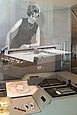 Der Graphomat Z64 ähnelt einem großen, grauen Tisch, auf dem ein Zeichenapparat montiert ist. Über den Tisch beugt sich die fotorealistische Pappfigur einer Frau. Das Bild der Frau ist schwarz-weiß. Sie trägt einen ärmelloses Kleid im Stil der Sechziger Jahre.