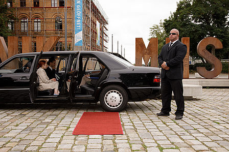 Schwarze Linmousine mit rotem Teppich. Ein Personenschützer steht neben der Limousine. Im Inneren sitzen drei Personen