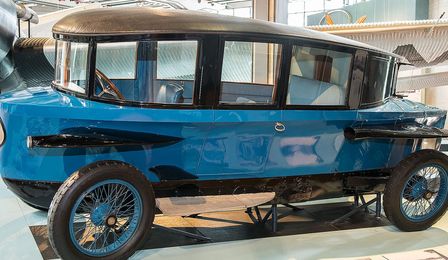 Ein historisches, längliches, blaues Fahrzeug, das durch seine aerodynamische, tropfenähnliche Form auffällt. Das Dach fällt nach hinten ab, Scheinwerfer und Schutzbleche an den Seiten erinnern an Flugzeugflügel.
