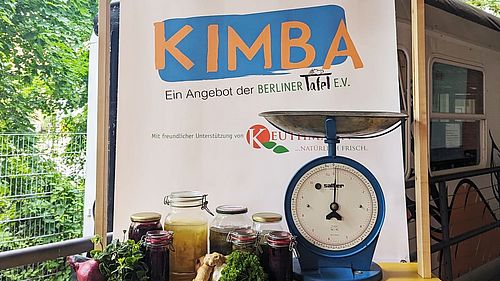 Vor dem KIMBA-Express steht eine Art Markstand mit einer blauen Gemüsewaage und verschiedenen Einweckgläseren mit eingelegtem Gemüse. 