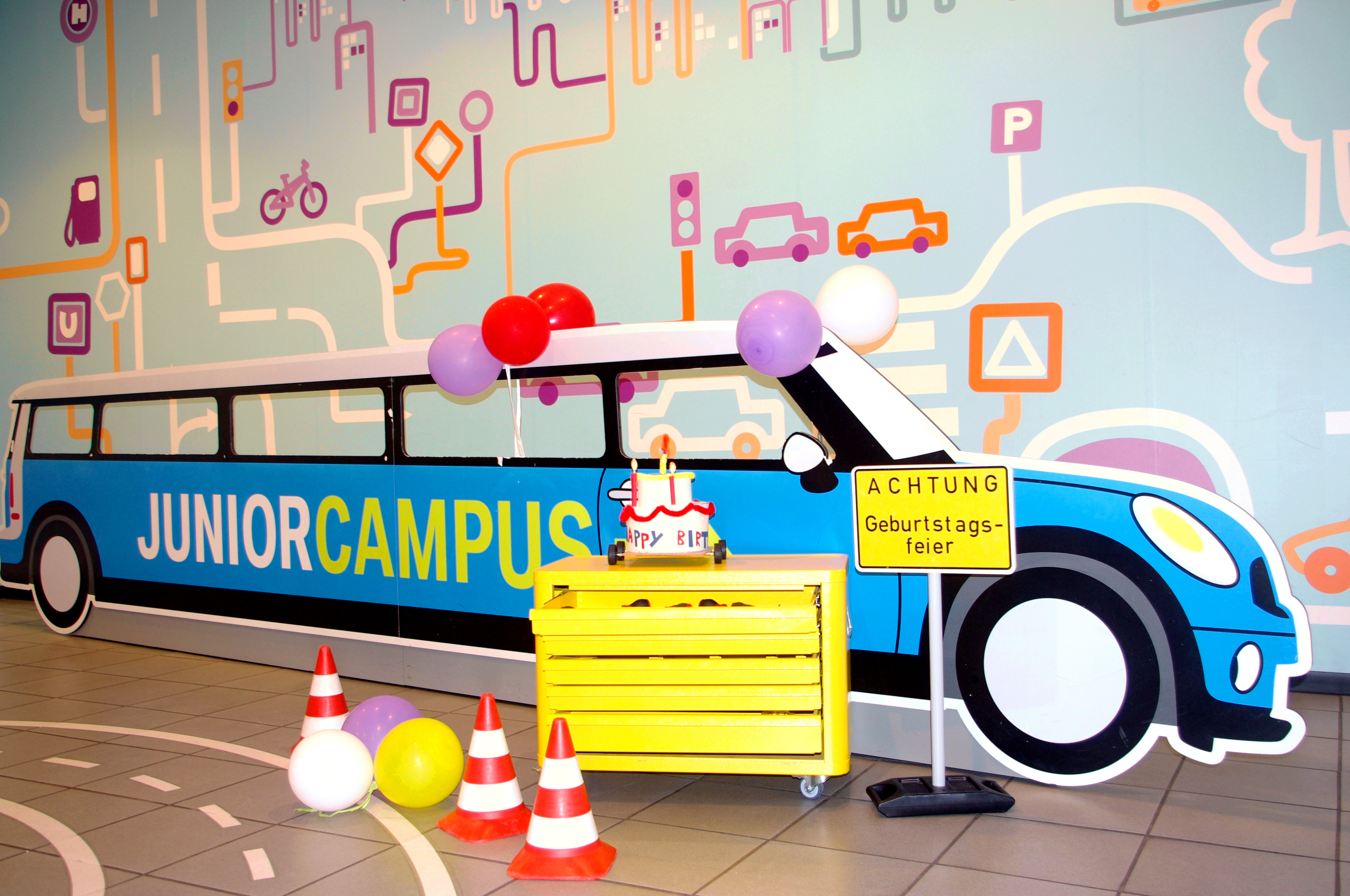 Ein etwa sechs Meter langer Aufsteller aus Pappe in Form eines Autos. Auf der Seite ist der Schriftzug „Junior Campus“ angebracht. Er ist mit Luftballons geschmückt. Davor steht ein Werkzeugschrank und ein Verkehrsschild mit der Aufschrift „Achtung Geburtstagsfeier“.