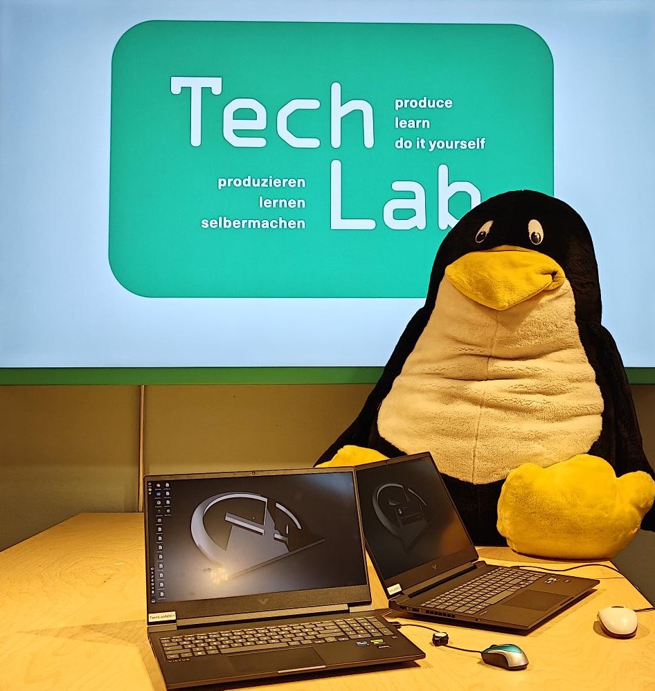 Ein Stoff-Pinguin sitzt auf einem Tisch, davor Elektroschrott.