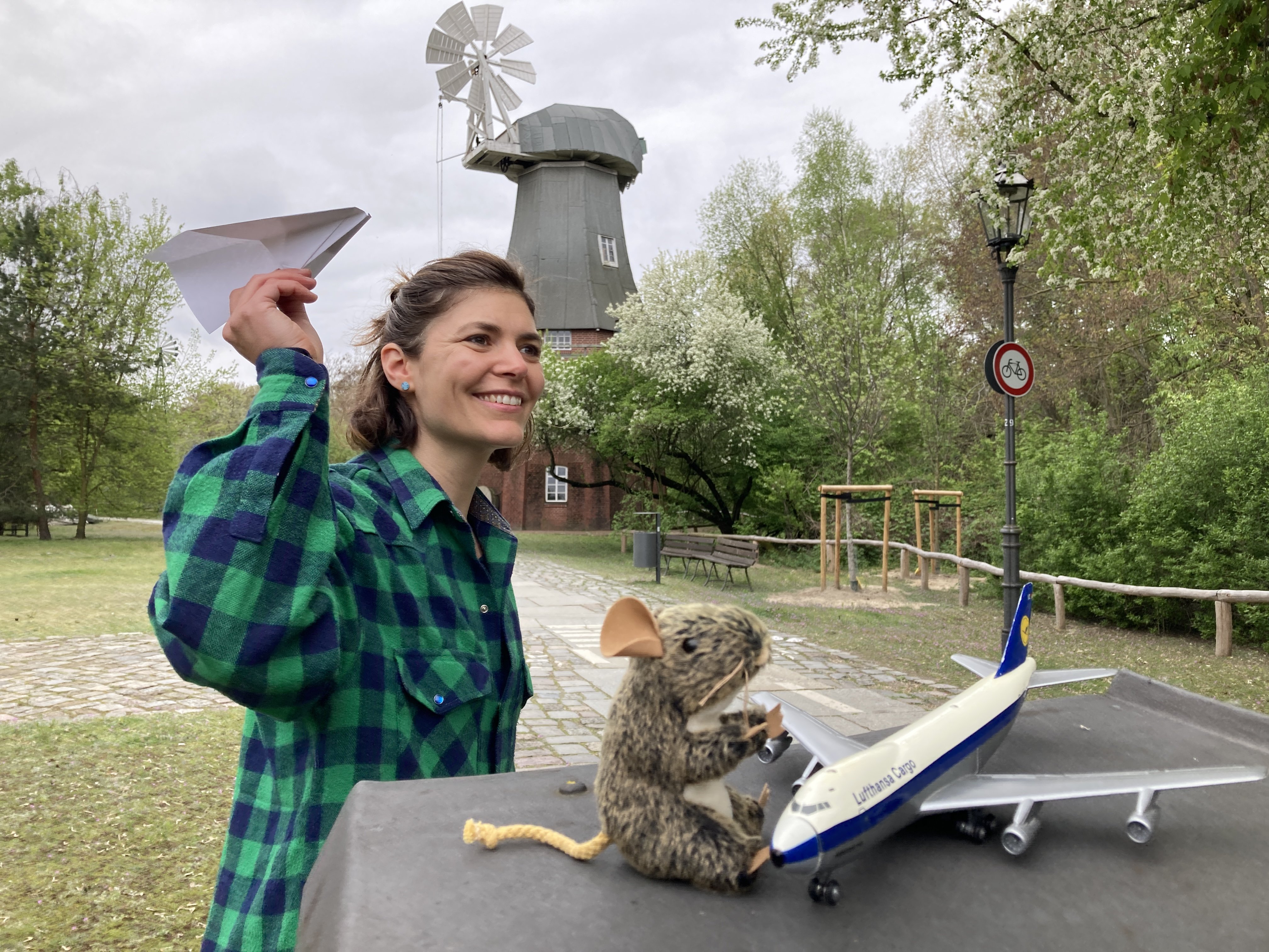 Im Museumspark. Im Hintergrund die Holländermühle. Im Vordergrund sitzt "Nanu" die Maus neben einem Modellflugzeug. Links neben ihr wirft eine Museumpädagoging ein Papierflugzeug. 