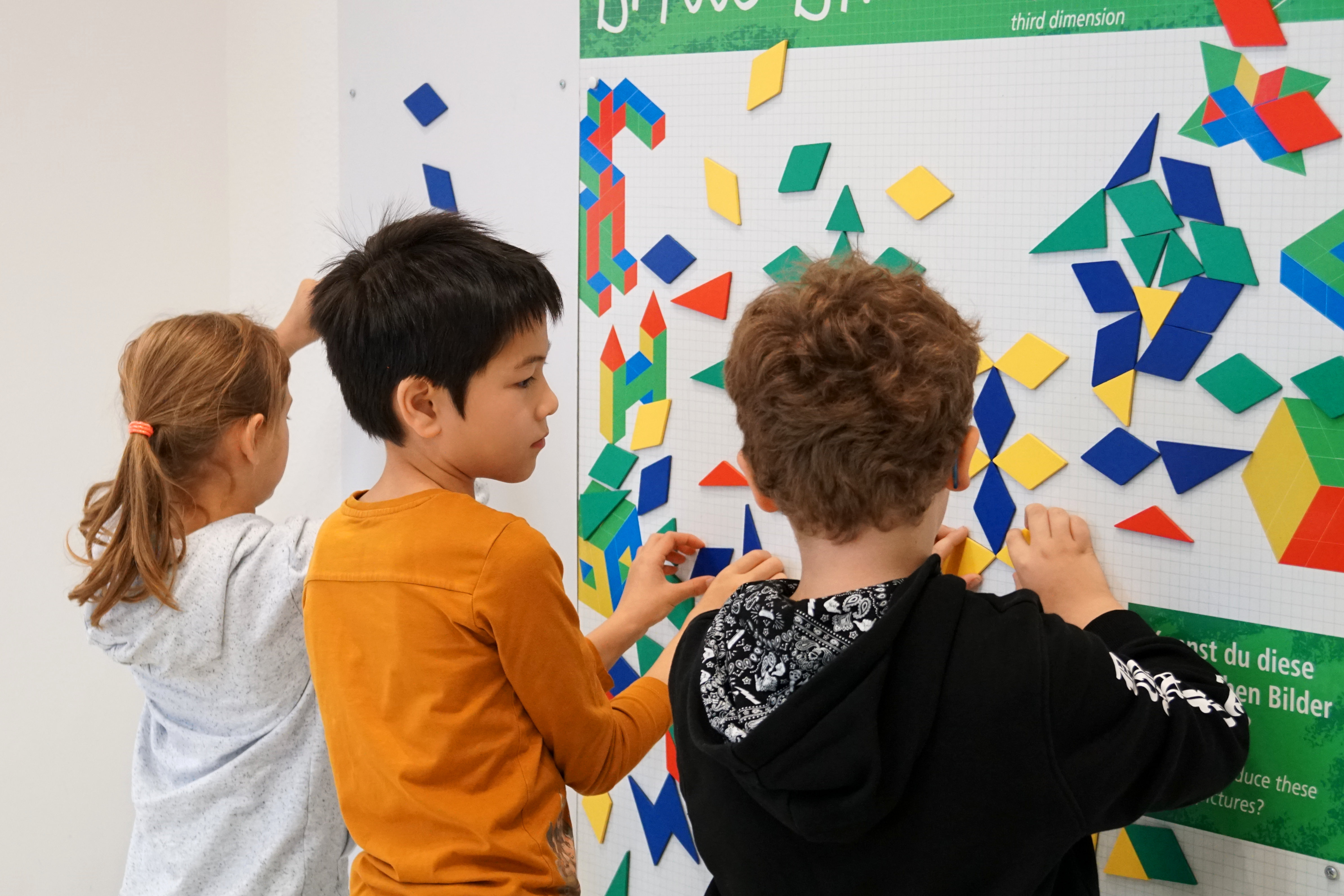 Drei Kinder stehen vor einer weißen Magnetwand und legen symmetrische Figuren aus verschiedenen bunten Formen.