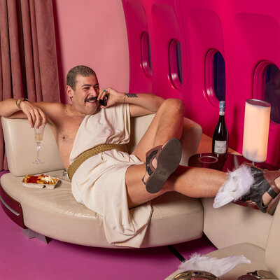 Das Farbfoto zeigt einen Mann mit Schnurrbart. Er liegt entspannt in einer luxuriösen Umgebung, die an das Innere eines Flugzeugs erinnert. Er trägt eine Art Toga mit goldenem Gürtel, an den Füßen Sandalen mit Flügeln. In einer Hand hält er ein Glas Sekt, in der anderen ein Telefon. Neben ihm stehen eine Pappschale mit Pommes Frites. Auf einem Sofa ihm gegenüber liegt ein Helm mit Flügeln. Die Szenerie ist in kräftigem Pink gehalten.