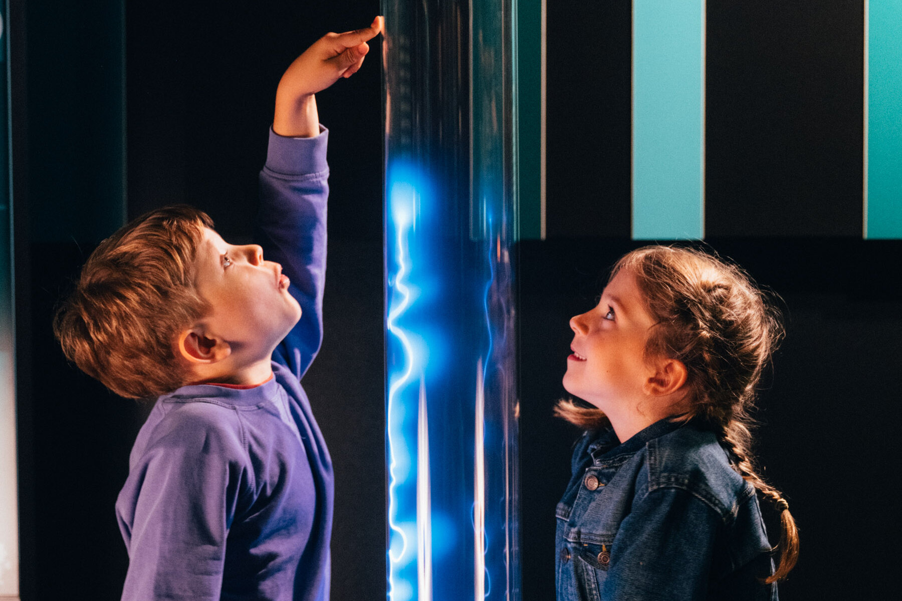 Zwei Kinder stehten in einem dunklen Raum um eine transparente Säule herum. Im Inneren der Säule leuchtet entlang der gesamten Höhe ein blauer Blitz. Eines der Kinder Kinder berührt die Plasmasäule mit seinen Händen.