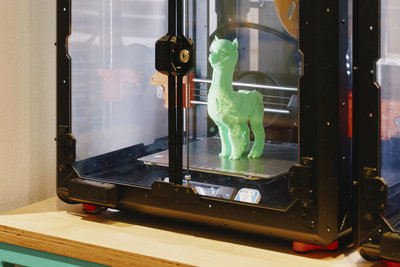 In einem schwarzen 3D-Drucker steht ein hellgrünes, gedrucktes Alpaka. Der Drucker steht auf einem Holztisch; rechts ist ein weiterer Drucker teilweise zu sehen.