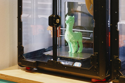 In einem schwarzen 3D-Drucker steht ein hellgrünes, gedrucktes Alpaka. Der Drucker steht auf einem Holztisch; rechts ist ein weiterer Drucker teilweise zu sehen.