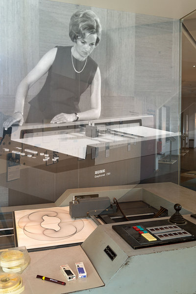 The Graphomat Z64 resembles a large, gray table with a drawing machine mounted on it. A photorealistic cardboard figure of a woman leans over the table. The image of the woman is in black and white. She is wearing a sleeveless dress in the style of the 1960s.