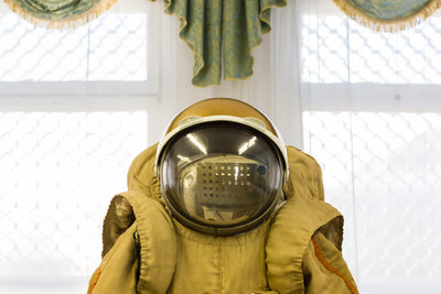 Das Bild zeigt zentral einen Kosmonautenanzug von der Taille aufwärts. Darauf sitzt ein kugelförmiger Helm mit großem Glasvisier. Im Hintergrund hängt über einem Fenster eine Zierschabracke mit Goldbordüre.